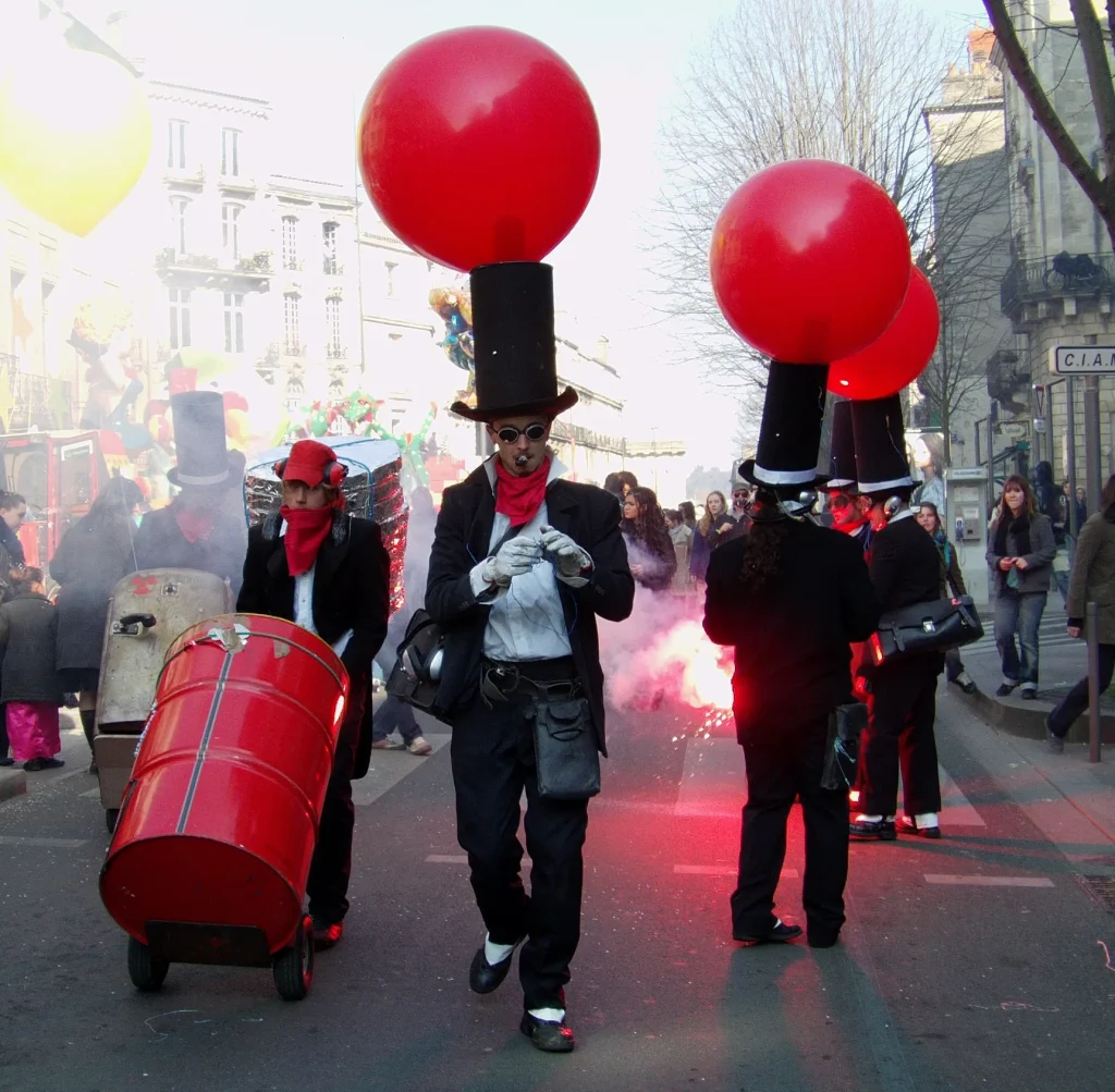 Une parade de rue : hauts-de-forme surmontés de ballons rouges, un bidon roulant