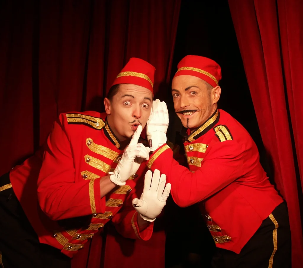 Les Grooms devant un rideau rouge, l'un fait signe de se taire